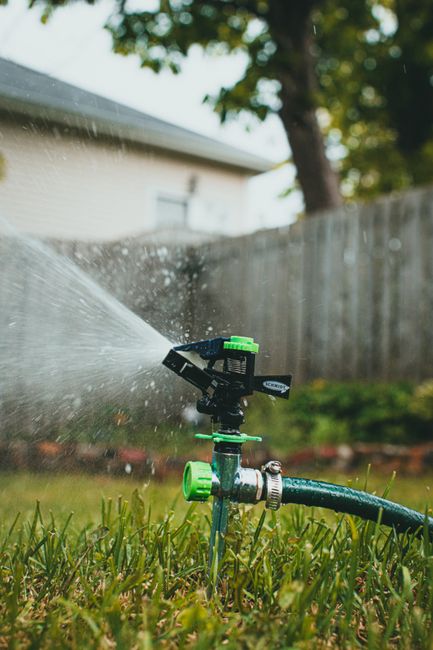 A garden scene during daytime featuring a well-maintained lawn with evenly cut grass and a garden bed bordered by bricks. In the foreground, a small, black irrigation sprinkler sprays water across the
